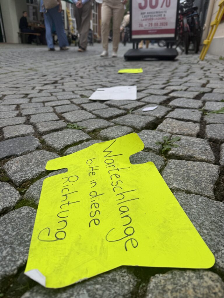 Handgeschriebenes Schild auf dem Pflaster: „Warteschlange bitte in diese Richtung“